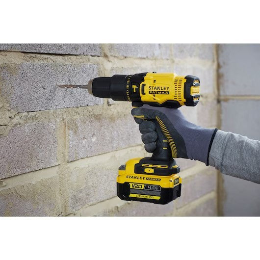 A yellow and black Stanley cordless drill being held by a gloved hand against a light gray brick wall. The drill bit is engaged with the masonry, and the Stanley branding is clearly visible on both the drill body and the battery pack.
