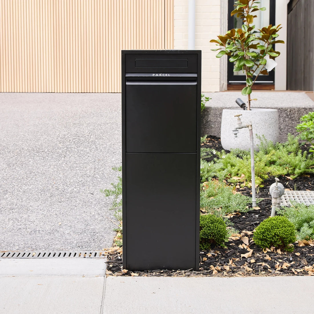 Zip Parcel Letterbox in black installed streetside beside a driveway and garden bed at a home entrance.