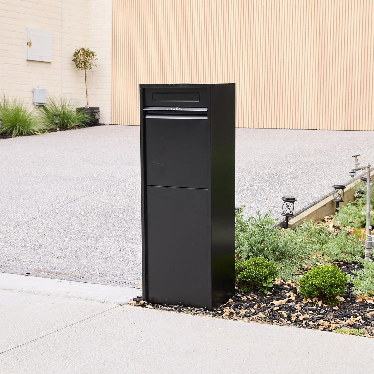 Zip Parcel Letterbox in black shown from a closer front angle beside a gravel driveway and low garden bed.